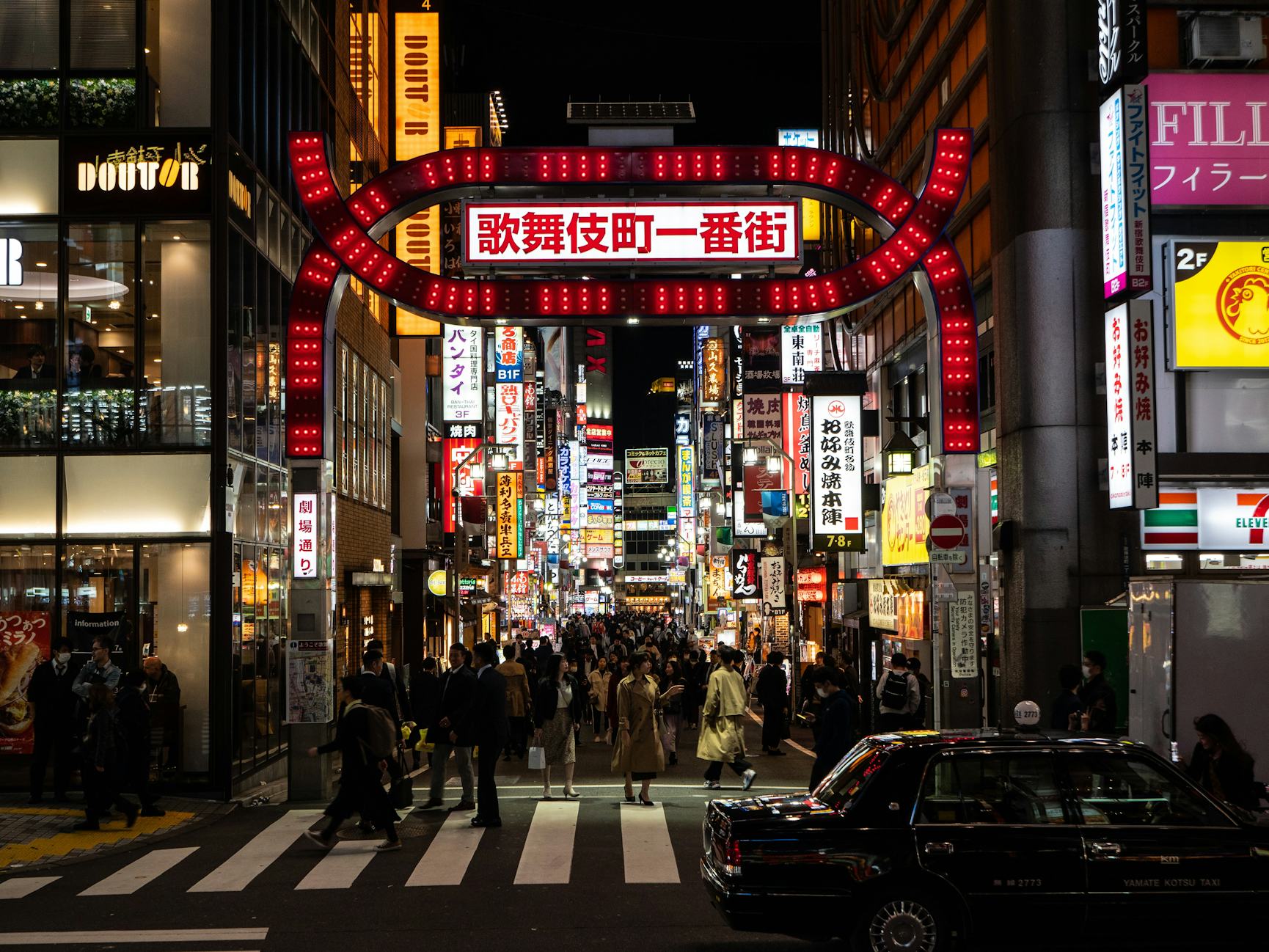 Night time image of street with crosswalk, crowds of people and lots of neon lights and bright billboards.