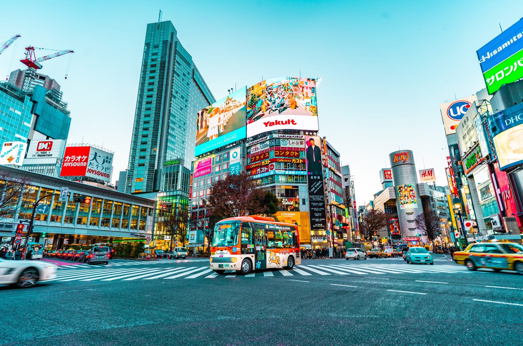 Japanese intersection with crossings and bus in centre. Bright billboards and cars all around.